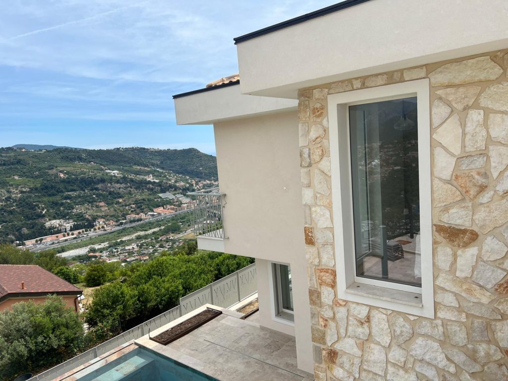 Vista esterna della villa ristrutturata a Sanremo: facciata con rivestimento in pietra naturale, serramenti in legno Campesato, terrazza con piscina a sfioro e panorama sulla vallata ligure
