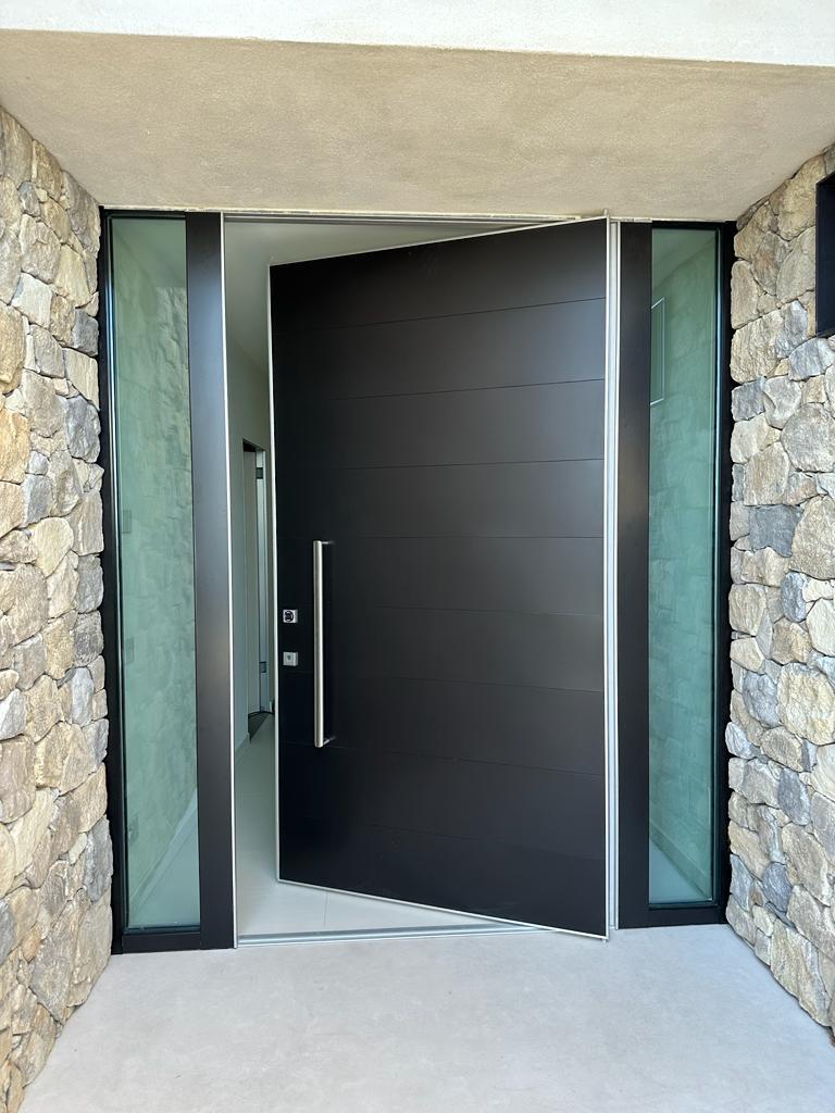 Porta blindata a bilico Oikos Synua installata in una villa moderna a Sanremo, con finitura nera opaca, maniglione in acciaio e fianchi luce vetrati laterali su parete in pietra a spacco