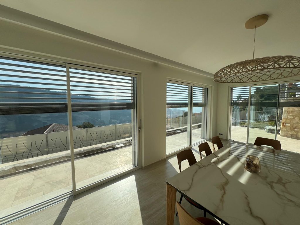 Soggiorno della villa a Sanremo con tavolo in marmo, lampadario di design, grandi vetrate scorrevoli in legno con frangisole Griesser e vista panoramica sulla terrazza e la Riviera Ligure