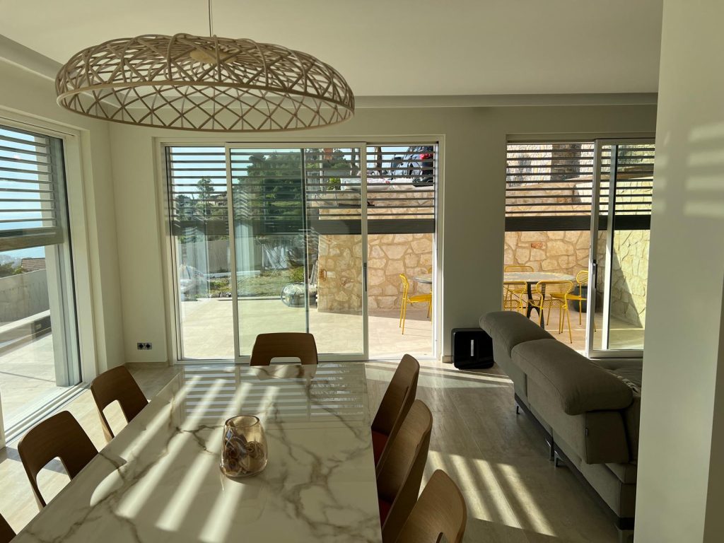 Interno del soggiorno arredato della villa a Sanremo: tavolo in marmo con sedie di design, lampadario intrecciato, divano e grandi vetrate scorrevoli in legno con vista sulla terrazza e il muro in pietra naturale